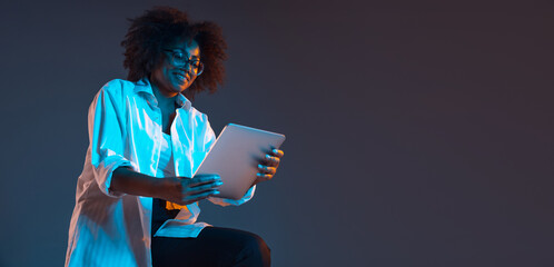 Portrait of African young girl in white shirt using tablet, gadget isolated on dark blue studio background. Concept of human emotions, facial expression, youth, sales, ad.