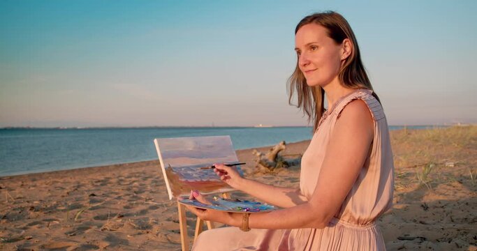 A woman sits on the sandy shore of the bay near an easel with a canvas and paints a picture with oil paints.