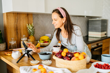 Middle age slim woman preparing food recipe with fresh fruits while streaming online with mobile smartphone cam for web kitchen masterclass channel.