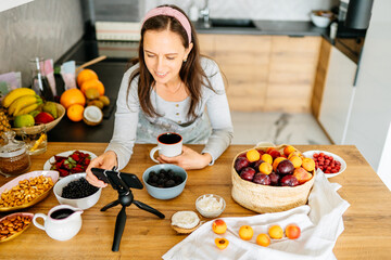 Congratulations holiday at distance, online call, video food blog at home. Happy pretty mature woman gesturing and look at smartphone webcam in kitchen interior with bright fresh fruits and berries.
