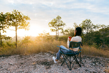 Fototapeta premium Rear view of happy young woman sitting on outdoor chair near a tent in the mountains, drinking coffee with sunrise in morning.
