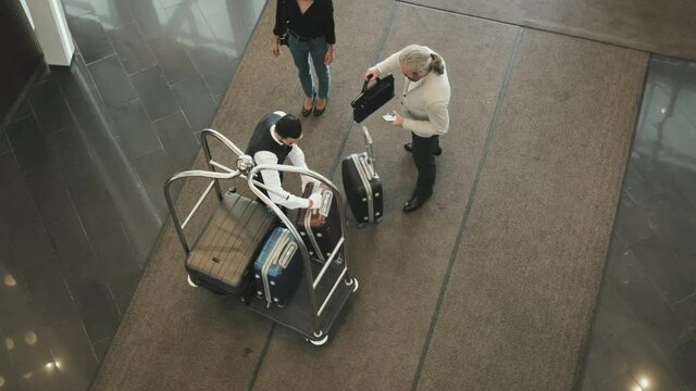 Top-view shot of modern tourist couple arriving at high-class hotel giving their baggage to young male porter placing it on cart