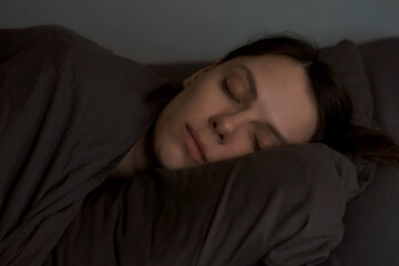Portrait of young woman is sleeping in the dark on pillow covering by blanket. Brown bed linen. Female's face with closed eyes. Resting and relaxing after busy day. Strong healthy sleep.