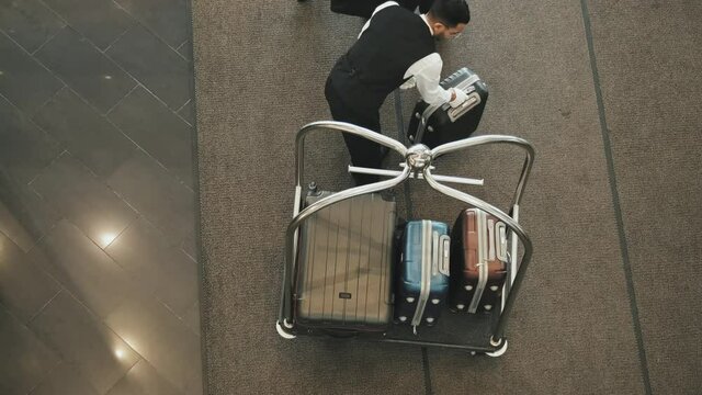 Top-view shot of contemporary tourist couple giving their suitcases to young male porter placing them on luggage cart in high-class hotel lobby