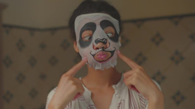 Funny 20s African American woman wearing funny panda cosmetics facial mask in bathroom looking at camera. Cheerful woman enjoying beauty procedures at home.