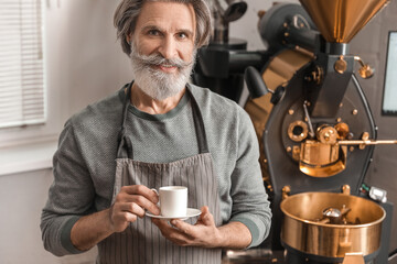 Senior man with cup of hot beverage near modern coffee roaster