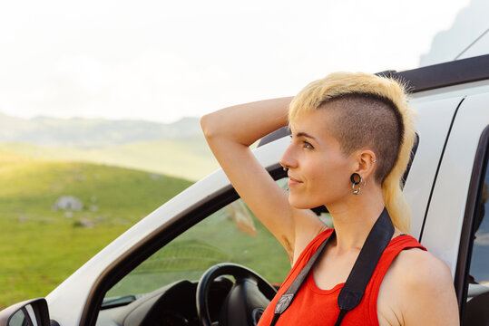 non-binary gender person in a red T-shirt and mohawk hair, with piercings, leaning on his car thinking in the midst of nature. - Powered by Adobe