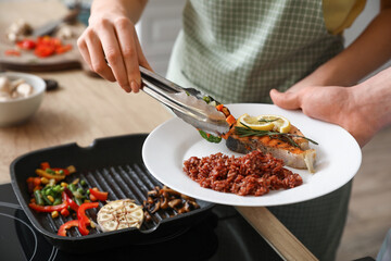 Woman putting tasty vegetables on plate with fish and rice in kitchen