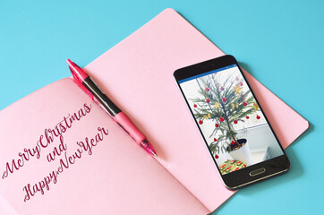 Smartphone with a christmas tree on display and pink notebook on blue table. Mock up for design