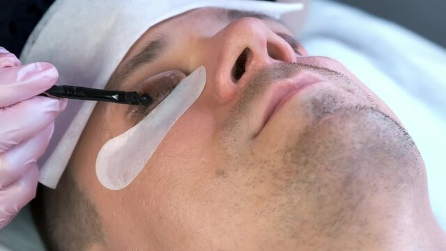 Cosmetologist puts black paint on the man's lashes using brush. Laminating eyelashes procedure in beauty salon. Beautician is painting lashes in black colour in cosmetology clinic, face closeup.
