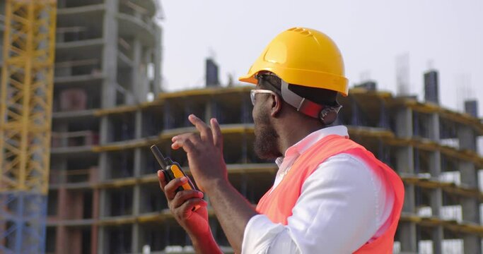 Builder in helmet is standing in front of the construction site, talking by walkie talkie control and communicate with worker on construction site. Contractor man using radio operation in industry.