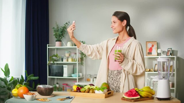 Young smiling woman making selfie photo holding glass with juice, food blogger recording video vlog on smartphone how to cook smoothie. Healthy sweet detox cocktail. 