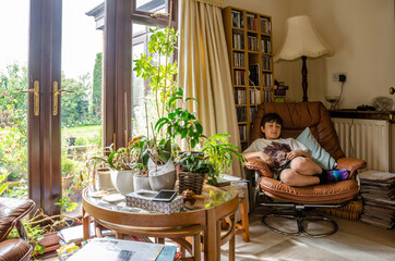 A boy relaxes in a comfy chair with a glass table covered with houseplants in front. A homely lifestyle scene from a family home.