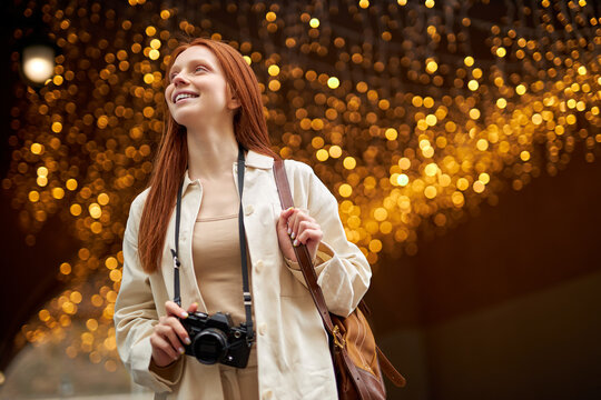 Relaxed friendly trendy young redhead woman with camera walk in city, boce garlands in the background. Portrait of beautiful lady in beige jacket enjoy walking alone outdoors. People lifestyle concept