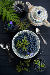 Vertical composition. Fresh blueberries on plates, spoons, blueberry branches and fern leaves, teapot on a dark gray wooden background. View from above