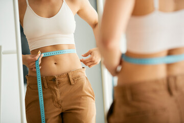 Reflection in a mirror of slim woman measures her waist with tape measure.