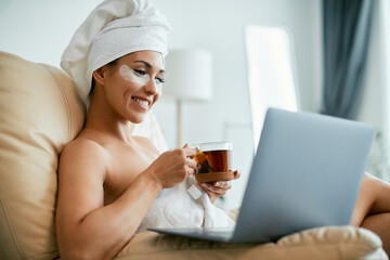 Young happy woman with  hydrogel under-eye recovery patches using laptop and drinking tea at home.