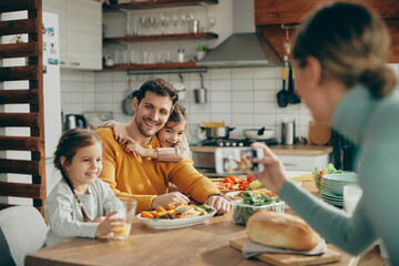 Happy father and daughters have fun while mother photographing them during a meal at dining table.