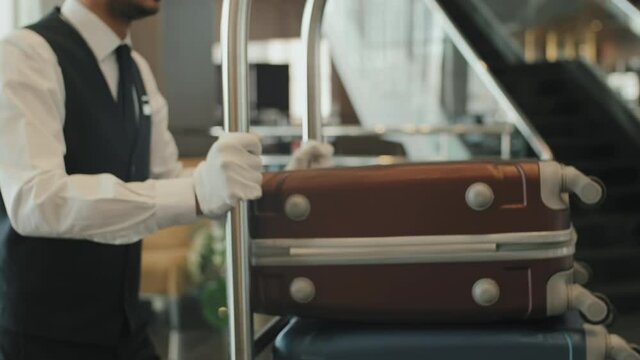 Midsection shot of unrecognizable male porter in uniform and gloves carrying baggage cart through modern hotel lobby