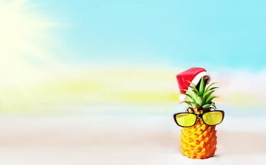 Attractive pineapple in stylish sunglasses on sand against sea water. Wearing christmas hat. Christmas and new year concept on a tropical beach.