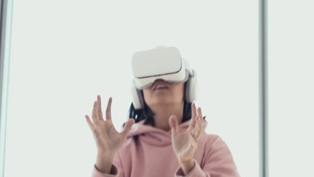 A girl is having fun in virtual reality glasses, standing against the background of a window and playing volleyball. High quality 4k footage