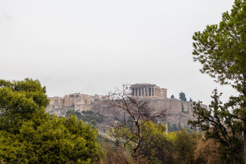 Fototapeta na wymiar Acropolis hill (Parthenon, Propylaea, Temples, Odeon of Herodes Atticus) view through green trees branches. Athens ancient historical landmark in city center from Filopappou Hill on cloudy day
