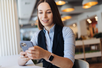 Young charming woman texting with smartphone while sitting alone in coffee shop, having conversation with mobile phone