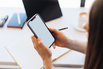 Female hands holding a smartphone with blank screen on working background, papers, laptop, white desk
