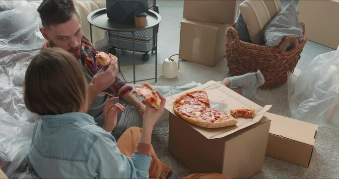 Cinematic move around young happy couple celebrating moving into new apartment eating pizza among cardboard boxes.