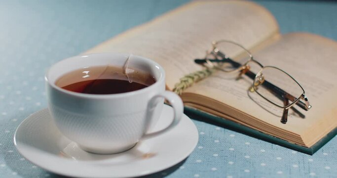 On the table is a white cup with a tea bag inside. Over a cup of tea, there is an open book and reading glasses.