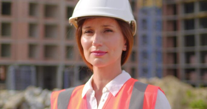 Portrait of Caucasian female industrial worker on construction site. Serious female industrial worker looking at camera wearing high vest and hard hat helmet. Front view. Close up.