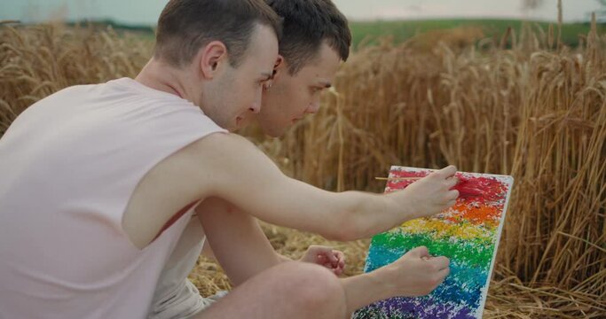 Two Romantic Teenage Gays sitting in the Field and Drawing Rainbow Picture. Using Wheat Stalk Focused on Drawing LGBT Sign. Couple of Men Lovers enjoys Time Together. Nature. Picnic. Artists.
