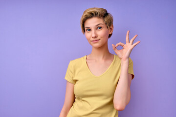 Short-haired Female showing ok gesture, everything will be OK. Isolated purple studio background. Young caucasian lady in casual yellow tshirt posing, smiling. Human emotions, people lifestyle