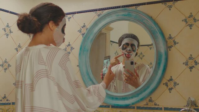 Millennial African American woman wearing funny panda cosmetics facial mask recording video on mobile phone standing in bathroom. Funny 20s woman making beauty procedures at home.