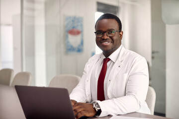 Happy African American doctor works on laptop and medical clinic and looking at camera.