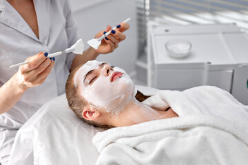Obraz na płótnie Canvas Cropped confident doctor cosmetologist or dermatologist making face mask in cosmetology office. Professional Beautician applying face mask on caucasian woman face lying on bed in bathrobe. copy space