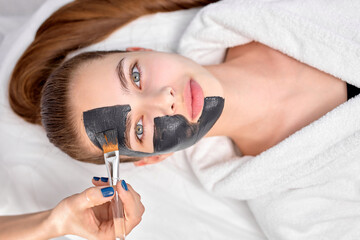 Woman relaxing with charcoal facial mask, cropped professional beautician applying on face of female client, using brush. Top view on female looking at camera, lying on bed in white bathrobe. beauty