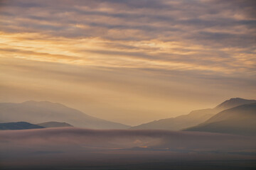 Scenic dawn mountain landscape with golden low clouds in valley among mountains silhouettes under cloudy sky. Vivid sunset or sunrise scenery with low clouds in mountain valley in illuminating color.