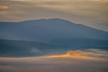 Scenic dawn mountain landscape with golden low clouds in valley among blue mountains silhouettes under cloudy sky. Sunset or sunrise scenery with low clouds in mountain valley in illuminating color.