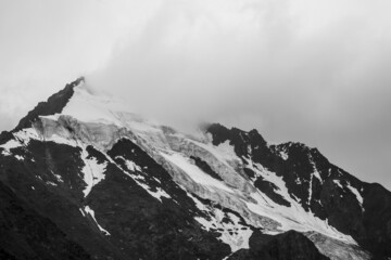 Fototapeta na wymiar Monochrome atmospheric mountains landscape with big snowy mountain top in low clouds. Awesome minimal scenery with white glacier on black rocks. High mountain pinnacle with snow in clouds in grayscale