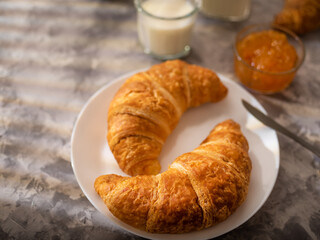 Breakfast. Two croissants with a golden brown crust on a white plate, orange jam, a glass of milk. Restaurant, cafe, hotel, home cooking. Advertising business. Low angle view.