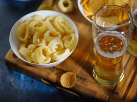 A glass of light beer, onion rings, snacks, chips on a wooden board. Dark blue background. The concept of rest in the company of friends, watching your favorite movie, sports programs, family vacation