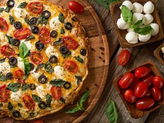 Pizza on a wooden tray and ingredients - tomatoes and mushrooms, mint leaves. High angle view. Wooden table. Restaurant, pizzeria, home cooking. Close-up. © Anton