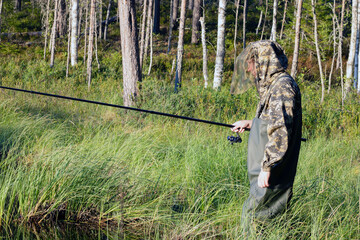 A fisherman in a camouflage suit in high boots in the water with a fishing rod in his hand, fishing in the lake.