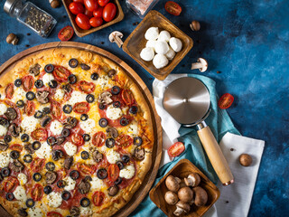 On a dark blue background, a large pizza on a wooden plate with many ingredients. Bright composition. High angle view. Restaurant and hotel business, pizzeria.