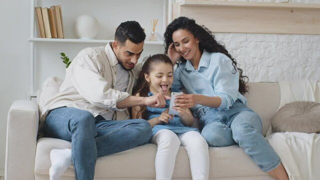 Spanish family sitting on couch using phone to take photos and record video. Parents with little daughter girl having fun trying funny masks online mobile application laughing child showing tongue