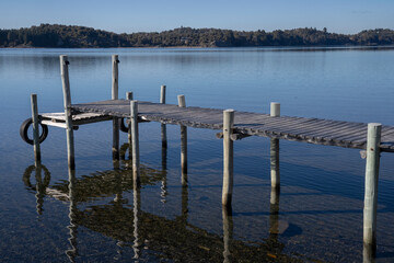 The wooden docks in the calm lake. The clear blue sky and pier reflected in the water surface. The forest and mountains in the background. 