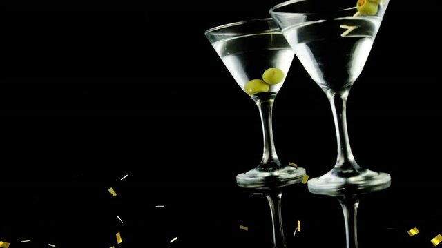 Golden confetti falling over olives in two cocktail glasses against black background