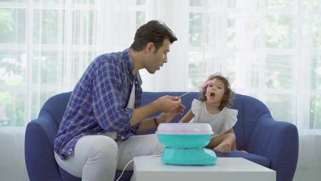 Hand father rolling cotton candy in candy floss machine and eat cotton candy together with daughter on sofa at new house
