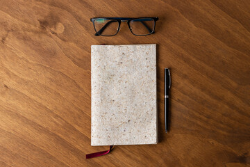 notebook of recycled paper of vegetal material and eyeglasses and a pen on wooden desk